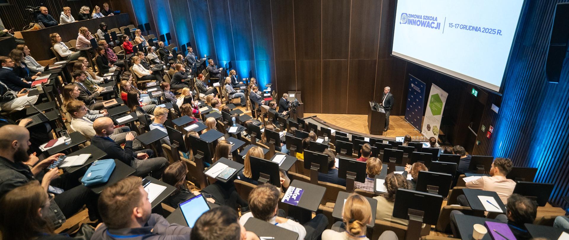 Zdjęcie z góry, widok na wielką aulę wypełnioną ludźmi, przed nimi na środku za mównicą stoi mężczyzna w garniturze i mówi do mikrofonu, za nim ekran z napisem Zimowa Szkoła Innowacji.