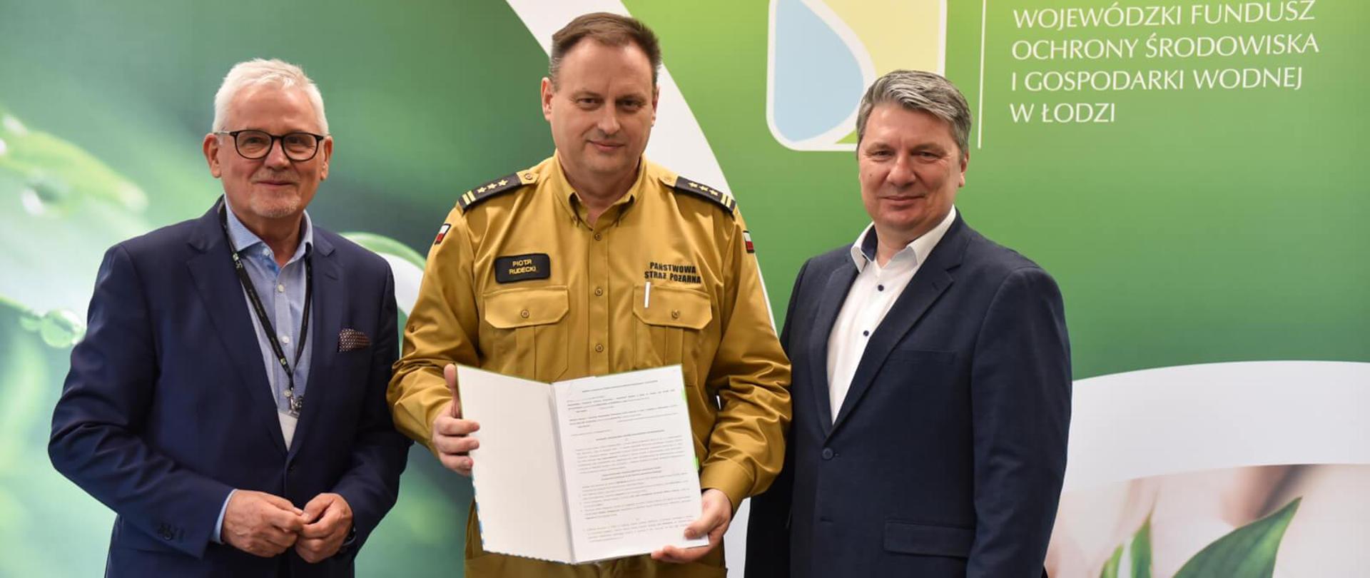 strażak i dwóch mężczyzn po bokach, strażak prezentuje podpisana umowę