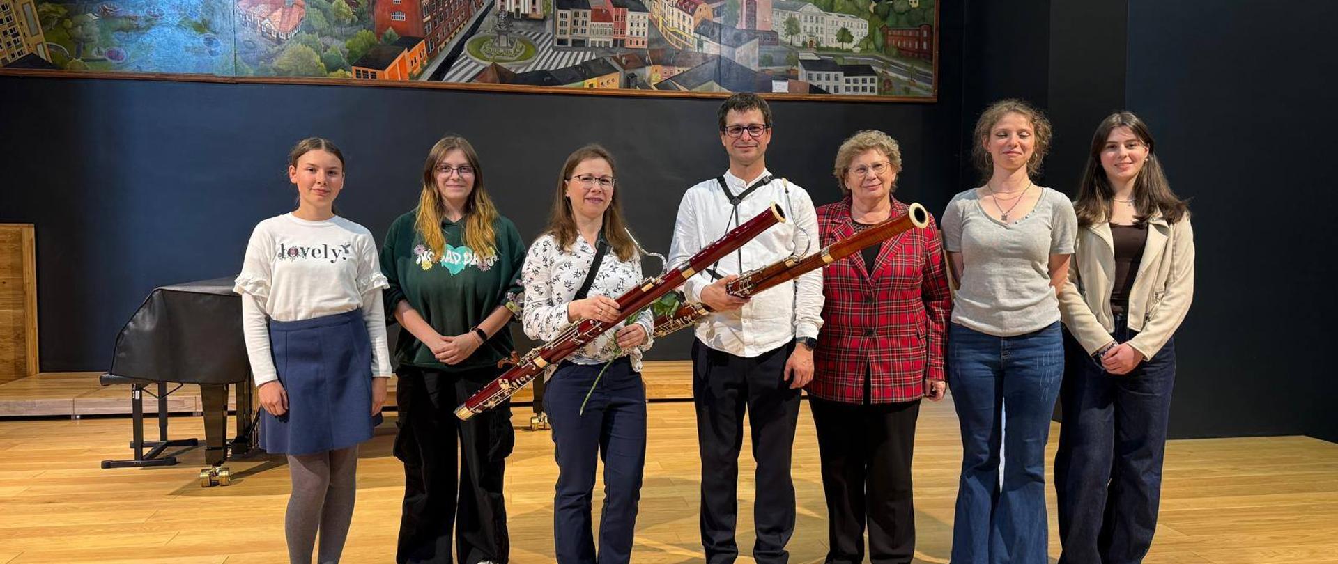 Kolorowe zdjęcie, na który stoją na scenie, w jednej linii, uczestnicy warsztatów fagotowych, nauczyciel klasy fagotu, dyrektor szkoły i prowadząca, na tle fortepianu i czarnej ściany z dużym kolorowym obrazem przedstawiającym najważniejsze budynki i miejsca w Rybniku. Prowadząca i nauczyciel klasy fagotu trzymają w rękach fagoty.