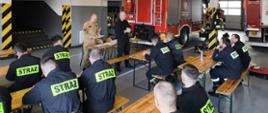 strażacy ubrani w czarne i piaskowe ubrania koszarowe siedzą przy ławkach. Ławki ustawione są na garażu, z tyłu widoczne samochody pożarnicze