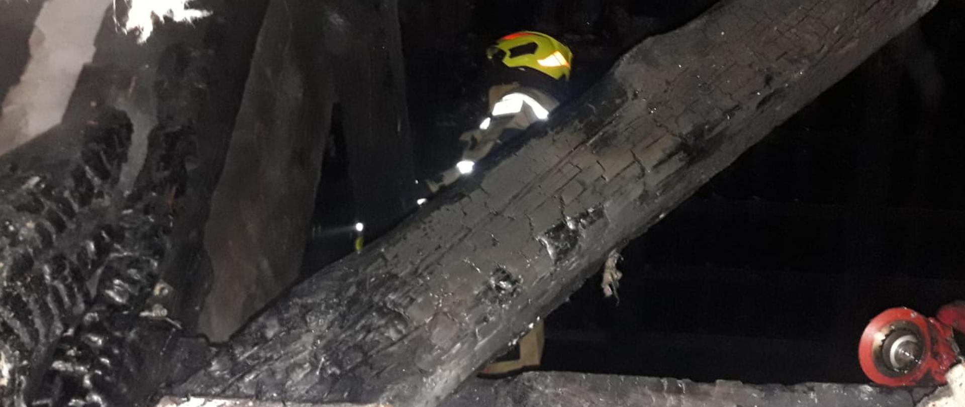 Na zdjęciu przepalona konstrukcja dachowa budynku mieszkalnego. W tle strażak sprawdzający czy nie ma zagrożenia. 