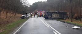 Widać leżący na drodze samochód dostawczy, po lewej na poboczu jest uszkodzony pojazd osobowy, w tle widać samochód strażacki i kilku strażaków