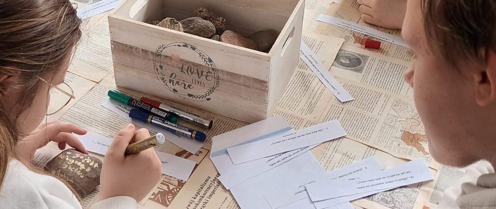 Pięcioro uczniów siedzi przy okrągłym stole, pracując nad zadaniem. Niektórzy z nich za pomocą kolorowych markerów piszą na gładkich kamieniach. Na środku stołu stoi drewniana skrzynka z napisem „Love lives here”, a obok leżą paski papieru z fragmentami tekstów oraz liczne pisaki. Stół przykryty jest papierowym obrusem z nadrukiem stylizowanym na stare gazety. W tle widać okno z widokiem na zieleń.