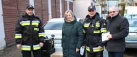 Obok sprzętu ratowniczego spakowanego w pudełkach stoi dwóch strażaków w czarno żółtych mundurach oraz kobieta i mężczyzna w zimowych ubraniach. Z tyłu zaparkowane samochody osobowe.