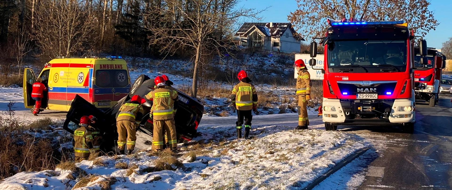 Jezdna i pobocze drogi, stojący strażacy przy przewróconych samochodzie, samochód strażacki i karetka pogotowia. W oddali stojące na jezdni samochody osobowe.