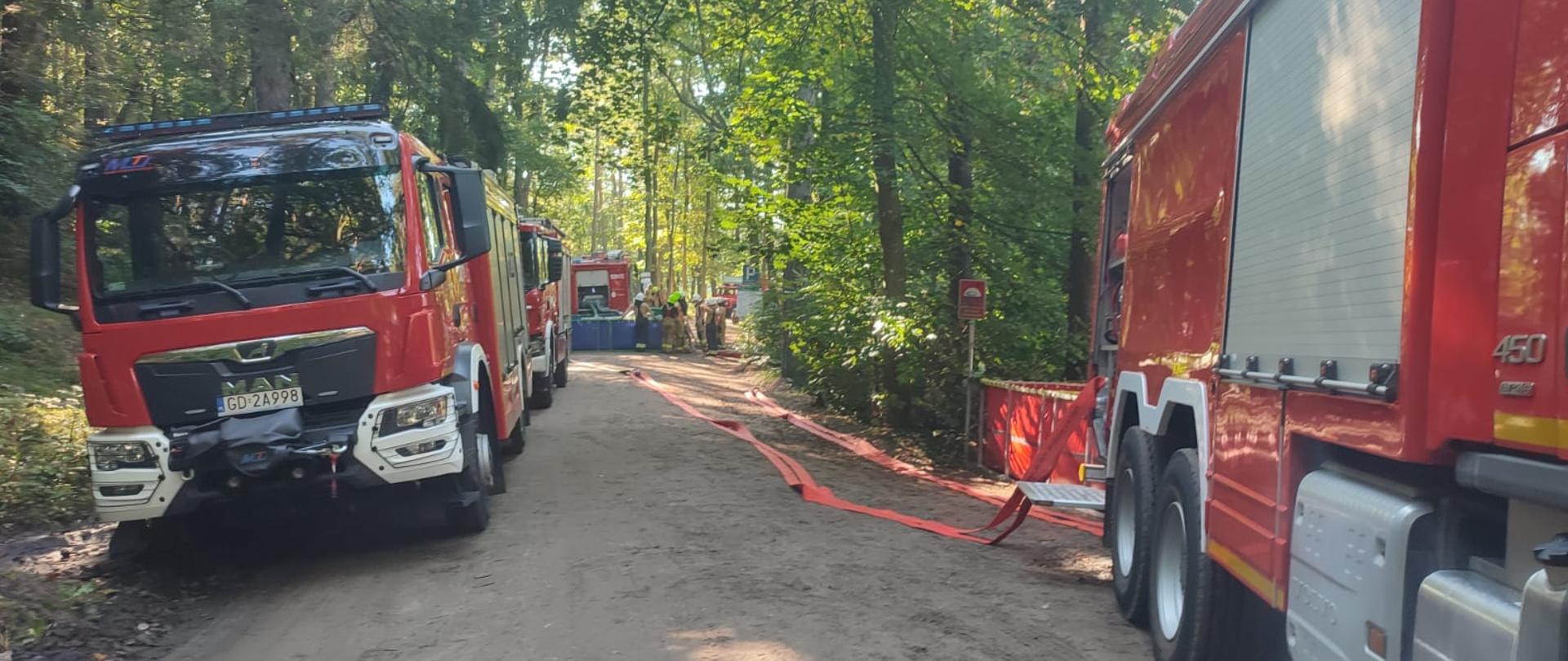 Droga Leśna. Po lewej i prawej stronie ustawione są wozy strażackie. Na drugim planie strażacy ustawiają zbiornik przenośny. Strażacy są ubrani w stroje bojowe.