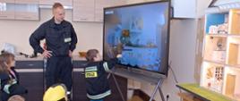 chłopiec zaznacza niebezpieczeństwo na obrazku wyświetlonym na interaktywnej tablicy reszta dzieci siedzi na dywanie prowadzący zajęcia strażak nadzoruje poczynania dzieci