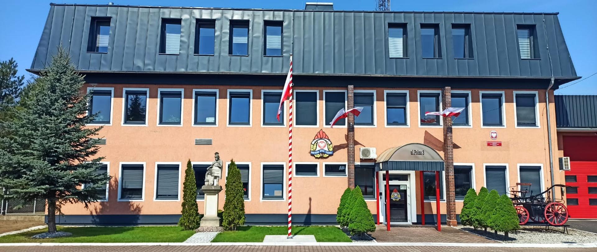 Zdjęcie przedstawia udekorowany obiekt komendy powiatowej PSP w barwy nbiło czerowne wraz z flagą na maszcie