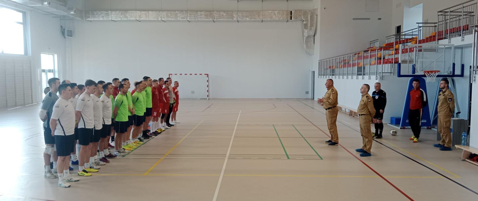 Widok na halę sportową. Po lewej stronie stoją drużyny trzech reprezentacji. Po prawej stronie naprzeciwko uczestników stoi czterech funkcjonariuszy PSP oraz sędzia główny.