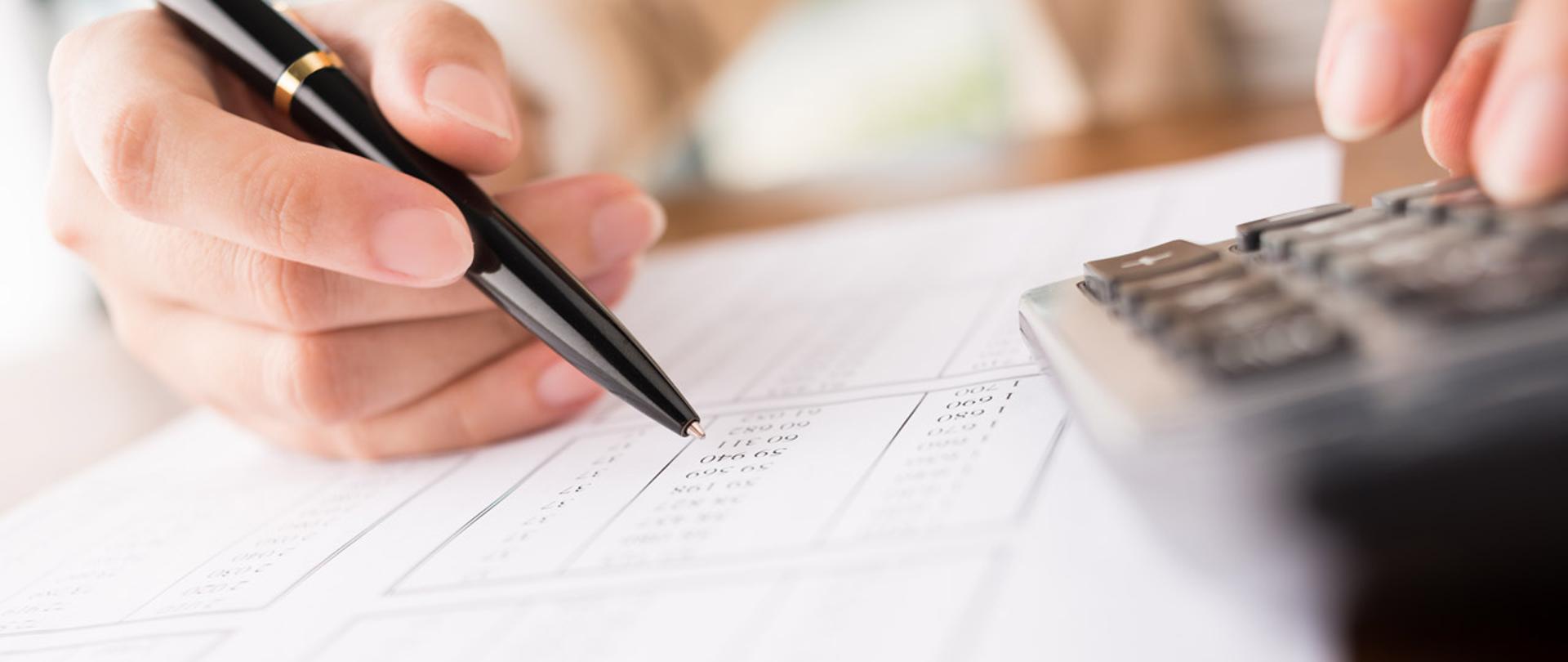 Osoba trzyma w rękach kalkulator i długopis. Kartka z liczbami