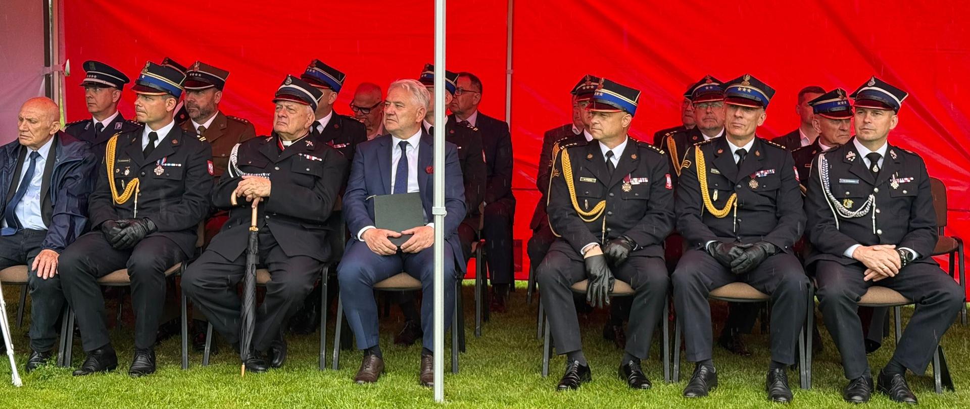 Zdjęcie przedstawia zaproszonych gości oraz wysokich rangą oficerów siedzących w rzędzie pod oficjalnym namiotem podczas uroczystości strażackiej. Zadaszenie stoiska: Nad siedzącymi rozciąga się intensywnie czerwony dach namiotu z dużym, białym, drukowanym napisem „STRAŻ POŻARNA”. Po prawej stronie widoczne jest logo Związku Ochotniczych Straży Pożarnych RP.Siedzący uczestnicy: W jednym rzędzie na krzesłach rozstawionych bezpośrednio na trawie siedzi kilkunastu mężczyzn. Większość z nich to oficerowie w ciemnogranatowych mundurach wyjściowych, czapkach rogatywkach ze złotymi galonami, w sznurach galowych i czarnych rękawiczkach. Ich piersi zdobią liczne baretki oraz medale.Goście cywilni: Wśród umundurowanych osób wyróżniają się dwaj mężczyźni w garniturach. Jeden (po lewej) ma na sobie ciemny garnitur i błękitną koszulę, drugi (bliżej środka) siedzi w niebieskim garniturze, białej koszuli i ciemnym krawacie, trzymając na kolanach ciemną teczkę z dokumentami.Konstrukcja: Przez środek kadru pionowo przebiega biała, metalowa rurka stelaża namiotu.