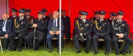 Zdjęcie przedstawia zaproszonych gości oraz wysokich rangą oficerów siedzących w rzędzie pod oficjalnym namiotem podczas uroczystości strażackiej. Zadaszenie stoiska: Nad siedzącymi rozciąga się intensywnie czerwony dach namiotu z dużym, białym, drukowanym napisem „STRAŻ POŻARNA”. Po prawej stronie widoczne jest logo Związku Ochotniczych Straży Pożarnych RP.Siedzący uczestnicy: W jednym rzędzie na krzesłach rozstawionych bezpośrednio na trawie siedzi kilkunastu mężczyzn. Większość z nich to oficerowie w ciemnogranatowych mundurach wyjściowych, czapkach rogatywkach ze złotymi galonami, w sznurach galowych i czarnych rękawiczkach. Ich piersi zdobią liczne baretki oraz medale.Goście cywilni: Wśród umundurowanych osób wyróżniają się dwaj mężczyźni w garniturach. Jeden (po lewej) ma na sobie ciemny garnitur i błękitną koszulę, drugi (bliżej środka) siedzi w niebieskim garniturze, białej koszuli i ciemnym krawacie, trzymając na kolanach ciemną teczkę z dokumentami.Konstrukcja: Przez środek kadru pionowo przebiega biała, metalowa rurka stelaża namiotu.