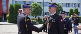 kilku strażaków ubranych w mundury galowe stoi na placu w szeregu, inny strażak podaje rękę w uścisku strażakowi obok którego stoi strażak ubrany w mundur galowy strażaka ochotniczej straży pożarnej, na dalszym planie stoją osoby cywilne i robią zdjęcia, w tle budynek komendy straży pożarnej, żywopłot oraz cztery rosnące drzewa, pogoda słoneczna