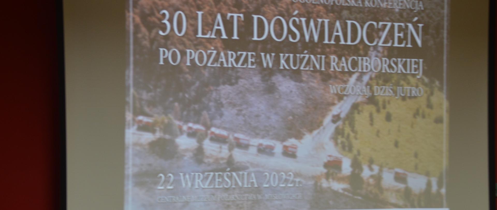 Konferencja upamiętniająca pożar lasu w Kuźni Raciborskiej. Świetlica Komendy Powiatowej PSP w Rawiczu. Ekran, na którym wyświetlany jest slajd tytułowy konferencji - na tle zdjęcia z pożaru umieszczony jest tytuł konferencji i data, a poniżej - logotypy współorganizatorów.