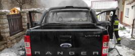 Centralnie na zdjęciu znajduje się czarny samochód terenowy marki Ford Ranger. Samochód otwarte ma drzwi i maskę od przedziału silnika. Za samochodem unosi się dym. z prawej strony zdjęcia stoi strażak z liną gaśniczą i gasi palący się silnik.