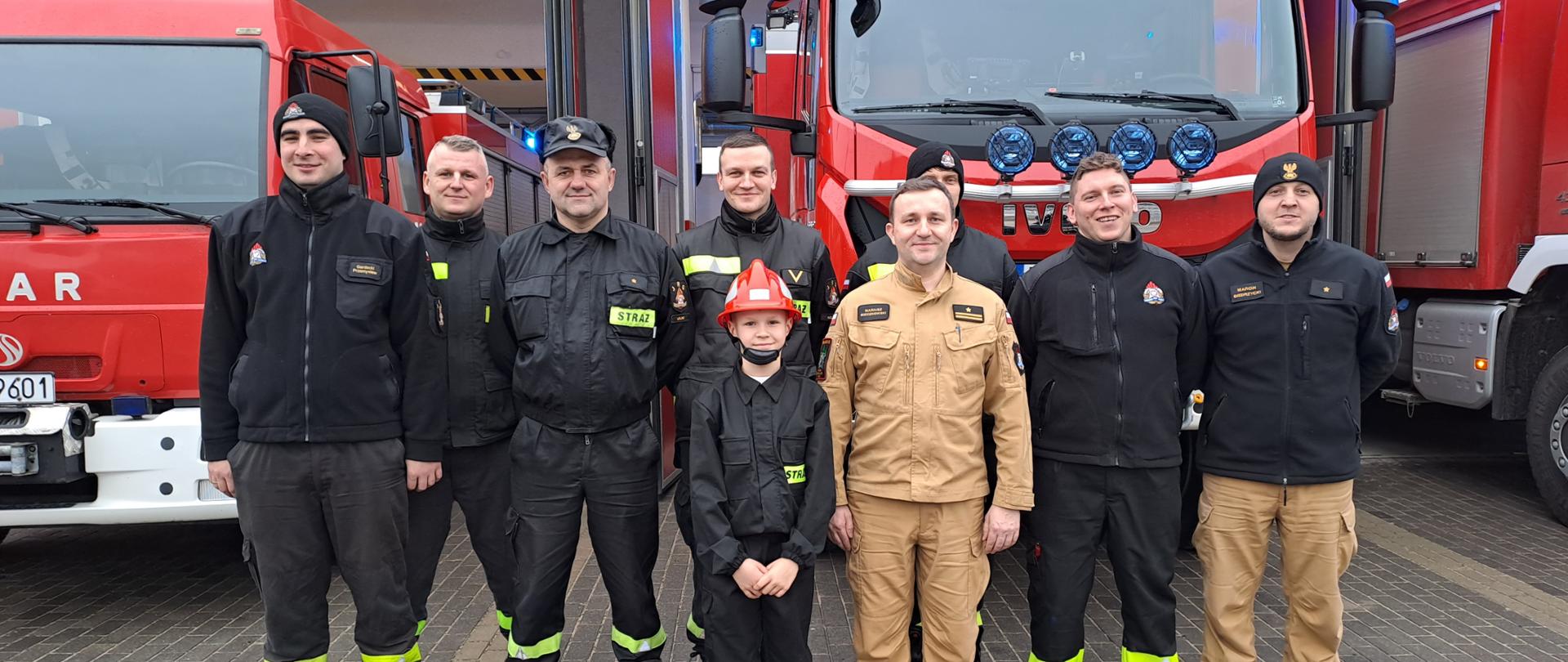 Widać strażaków stojących w szeregu na tle pojazdów pożarniczych.