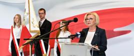 Wiceminister Machałek stoi za mównicą i mówi do mikrofonu, za nią troje elegancko ubranych uczniów ze sztandarem stoi na tle polskiej flagi.