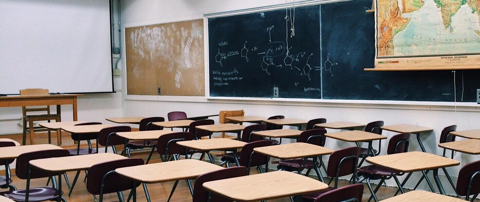 Pusta klasa szkolna z pojedynczymi ławkami i krzesłami w tle tablice ze wzorami chemicznymi i mapa świata