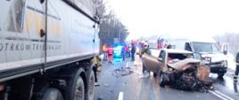 Zdjęcie przedstawia uszkodzony samochód osobowy. W tle widać Pogotowie Ratunkowe oraz Policję. Na drodze znajduje się dużo połamanych części samochodowych.