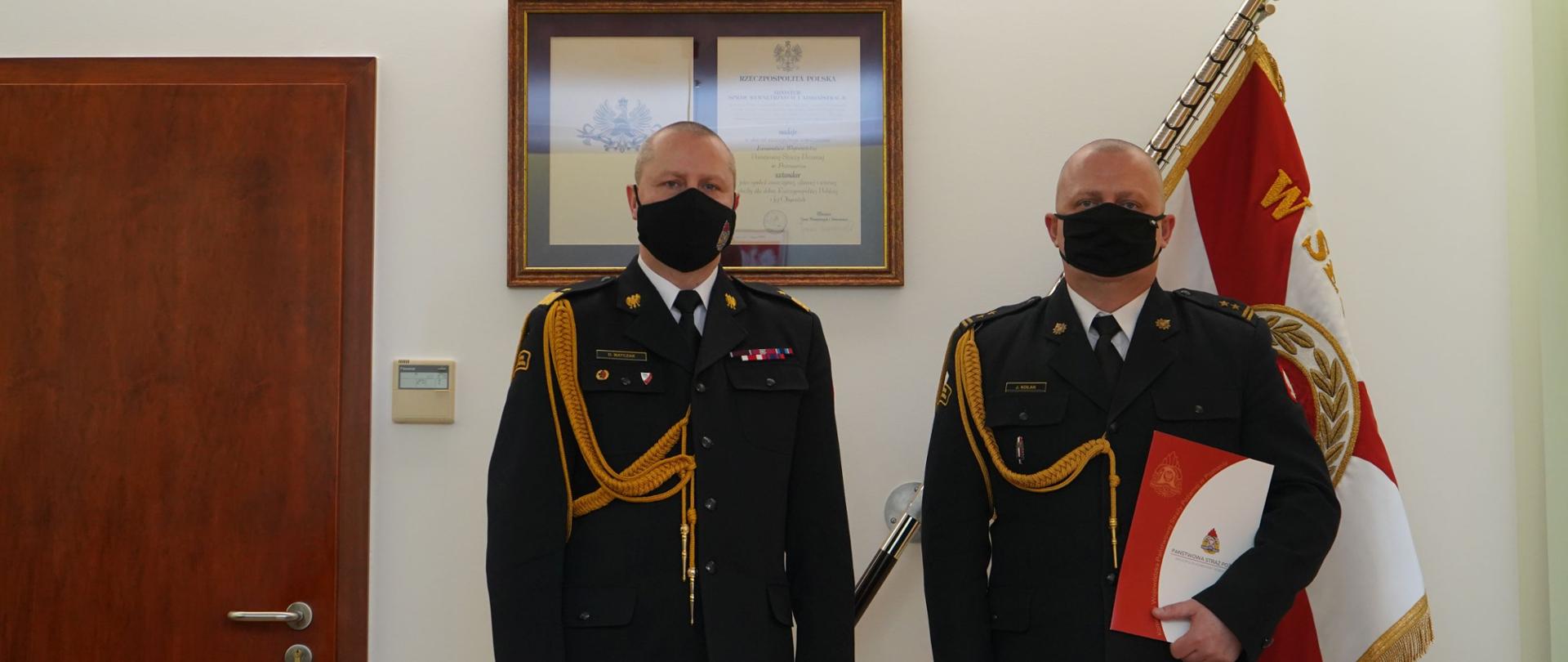 dwóch strażaków w maseczkach stoi na baczność, ubrani w w ciemne mundury i białe koszule, w tle sztandar