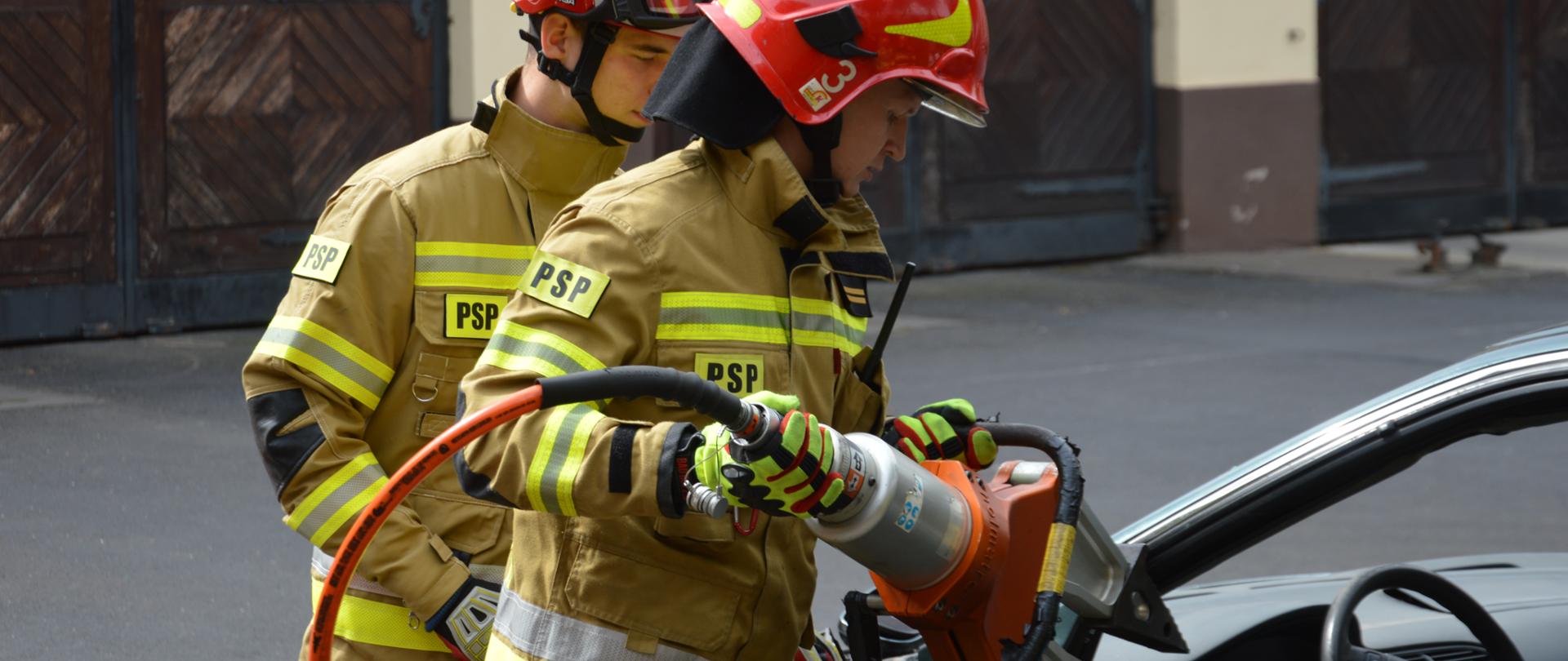 Na zdjęciu widocznych dwóch strażaków w pełnym umundurowaniu bojowym wykonujący ćwiczenie cięcia wraku pojazdu przy użyciu narzędzi hydraulicznych