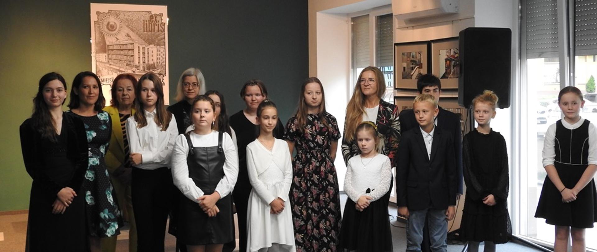 Zdjęcie przedstawia uczniów i nauczycieli stojących na tle zielonej ściany w lewej części zdjecia i na tle okien po prawej stronie zdjęcia