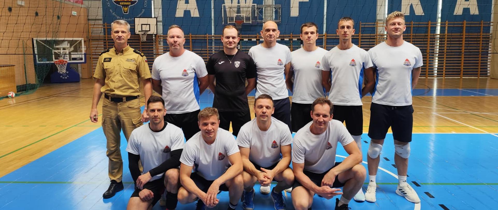Na zdjęciu jest 11 mężczyzn, 10 z nich ubranych w stroje sportowe sugerujące uprawianie sportu (grali w siatkówkę) Jeden z nich ubrany w mundur w kolorze piaskowym. Panowie stoją wewnątrz hali sportowej za nimi widać rozciągniętą siatkę co świadczy o tym iż grali w siatkówkę