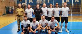 Na zdjęciu jest 11 mężczyzn, 10 z nich ubranych w stroje sportowe sugerujące uprawianie sportu (grali w siatkówkę) Jeden z nich ubrany w mundur w kolorze piaskowym. Panowie stoją wewnątrz hali sportowej za nimi widać rozciągniętą siatkę co świadczy o tym iż grali w siatkówkę