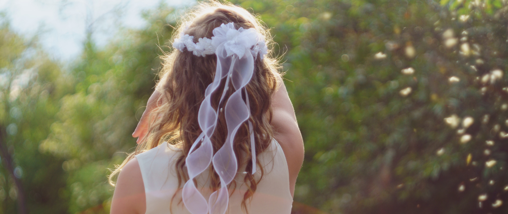 Odwrócona plecami dziewczynka pierwszokomunijna ubrana w biała sukienkę oraz mająca wianek na głowie.