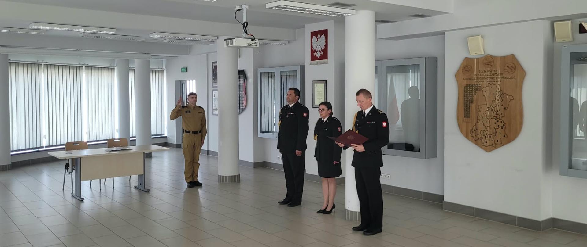 Fotografia ukazuje oficjalną część uroczystości w przestronnej, jasnej sali komendy. Po lewej stronie, tyłem/półprofilem do obiektywu, stoi nowo przyjęty strażak w piaskowym mundurze, z podniesioną do góry prawą dłonią w geście ślubowania (dwa palce uniesione do przysięgi). Przed nim, w centralnej części sali, stoi troje funkcjonariuszy kadry kierowniczej w ciemnych mundurach galowych (dwóch mężczyzn i jedna kobieta) – jeden z nich odczytuje rotę ślubowania z trzymanego w rękach dokumentu. Po prawej stronie, w głębi sali, stoi trzyosobowy poczet sztandarowy w pełnym umundurowaniu galowym i rogatywkach, trzymający sztandar jednostki, oddający honor nowemu koledze. Całość odbywa się w oficjalnej, ceremonialnej atmosferze.