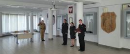 Uroczysta zbiórka w sali komendy PSP w Bochni. Po lewej stronie młody strażak w piaskowym mundurze składa przysięgę z uniesioną dłonią. Naprzeciw niego stoi troje mundurowych w ciemnych strojach galowych, z których jeden czyta tekst z czerwonej teczki. W tle widać godło Polski, drewnianą tablicę pamiątkową i gabloty na ścianach.