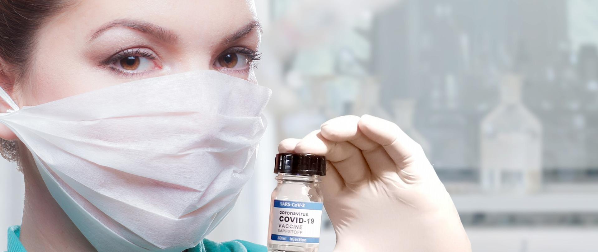 Kobieta w zielonym, jednorazowym fartuchu i maseczce ochronnej ba twarzy trzyma w dłoni osłoniętą rękawiczką jednorazową fiolkę ze szczepionka przeciw Civid-19