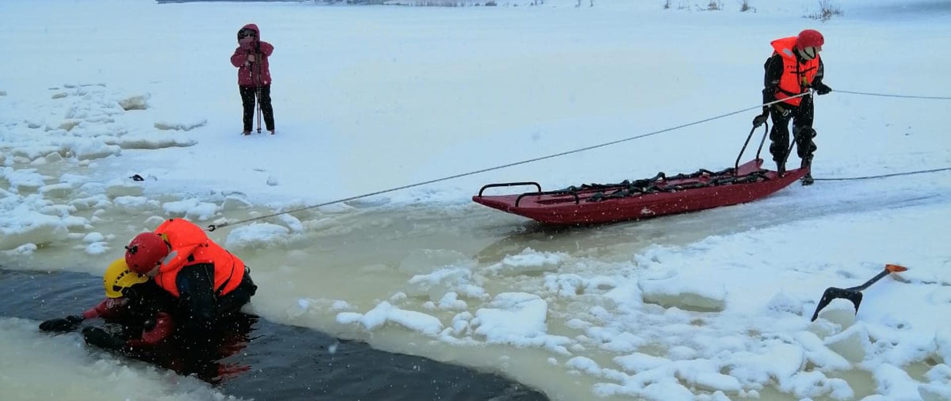 Zdjęcie przedstawia ratowanie osoby tonącej pod którą załamał się lód. W wodzie znajduje się jeden ze strażaków ubrany w specjalne ubranie ochronne do prowadzenia działań w wodzie. Przy nim znajduje się drugi strażak ubrany w ubranie ochronne z przywiązaną do niego linką ratowniczą, który próbuje go wyciągnąć z wody. Za nimi znajduje się kolejny ratownik ze specjalnymi saniami umożliwiającymi poruszanie się po zamarzniętych akwenach wodnych. 