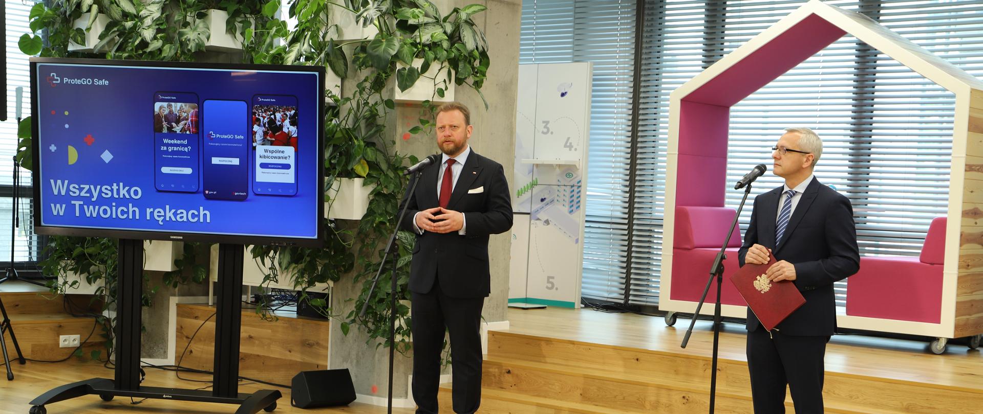 Zdjęcie z konferencji z udziałem Marka Zagórskiego, sekretarza stanu w KPRM oraz Łukasza Szumowskiego, byłego ministra zdrowia. Obok monitor z wyświetlonym hasłem: ProteGO Safe. Wszystko w Twoich rękach.