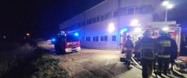 Jest pora nocna. Przy hali produkcyjnej stoją dwa samochody pożarnicze. Jeden z nich ma rozłożony maszt oświetleniowy. Przy samochodzie stoi grupka strażaków