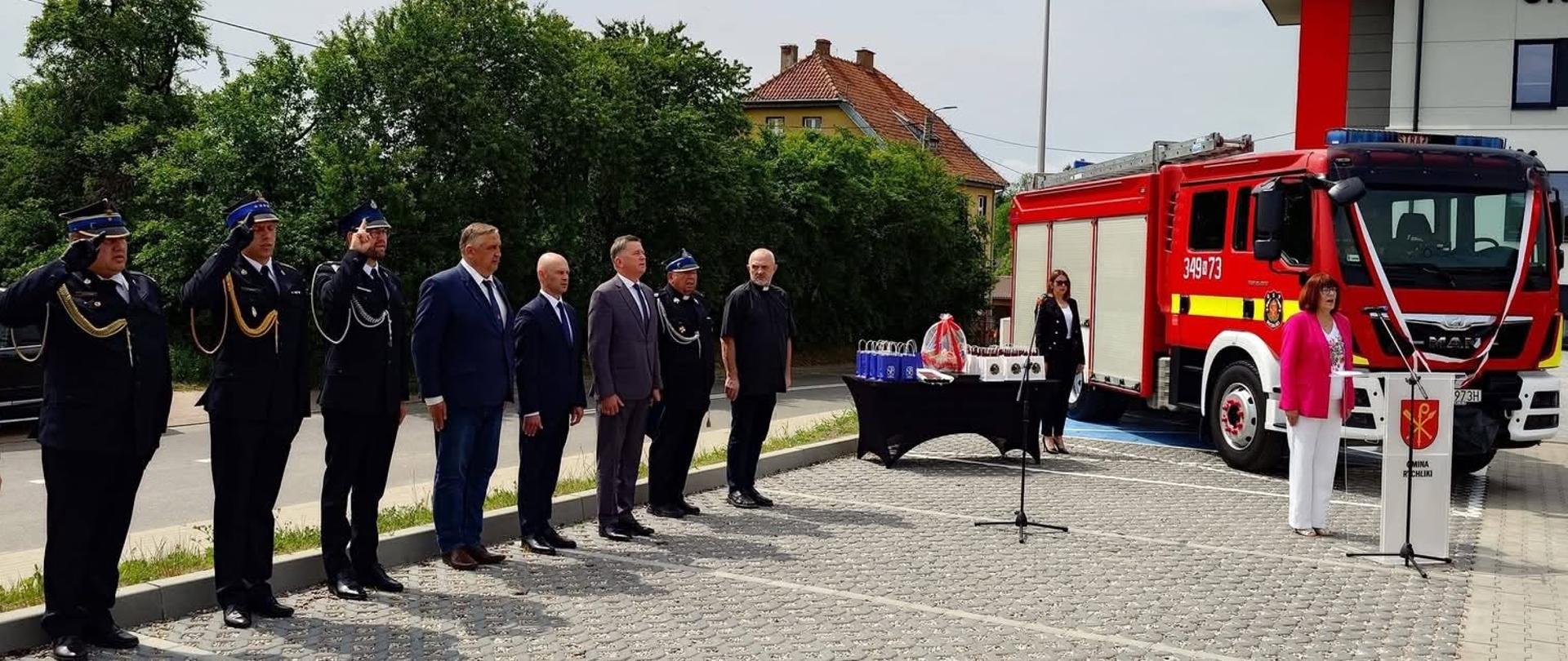 Przed budynkiem strażnicy odbywa się oficjalna uroczystość z udziałem strażaków i przedstawicieli samorządu. Na placu, w równym szyku, stoją funkcjonariusze w galowych mundurach, z których część oddaje salut. Obok ustawiono mównicę z herbem i napisem Urząd Gminy, a na stoliku przykrytym czarnym obrusem widoczne są przygotowane nagrody lub upominki. W tle stoi czerwony samochód pożarniczy, co podkreśla charakter wydarzenia związanego ze służbą pożarniczą.