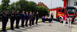 Przed budynkiem strażnicy odbywa się oficjalna uroczystość z udziałem strażaków i przedstawicieli samorządu. Na placu, w równym szyku, stoją funkcjonariusze w galowych mundurach, z których część oddaje salut. Obok ustawiono mównicę z herbem i napisem Urząd Gminy, a na stoliku przykrytym czarnym obrusem widoczne są przygotowane nagrody lub upominki. W tle stoi czerwony samochód pożarniczy, co podkreśla charakter wydarzenia związanego ze służbą pożarniczą.
