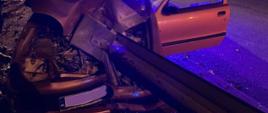 Rozbity samochód osobowy, który zatrzymał się na przydrożnej barierze. W tle strażacy, radiowóz policyjny i samochód gaśniczy.