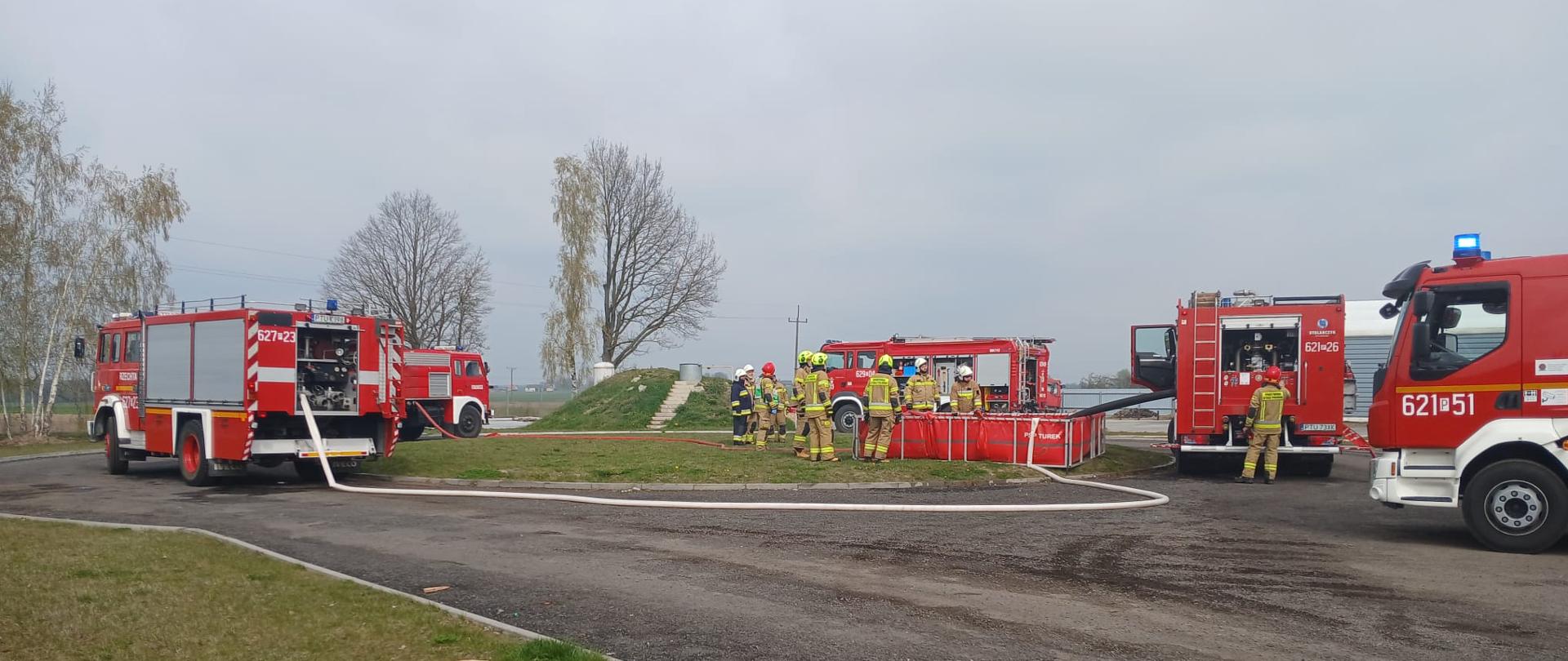 Kilka czerwonych wozów strażackich stoi na placu, a strażacy w ubraniach ochronnych obsługują sprzęt i zbiornik z wodą podczas ćwiczeń na otwartym terenie.