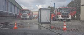 Teren ćwiczeń ratowników, przedstawia pojazdy pożarnicze, węże pożarnicze napełnione wodą i podłączone do rozstawionej kabiny dekontaminacyjnej oraz kurtyny wodnej