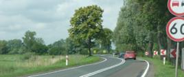 Kręta droga wśród drzew i łąk. Dwa ostre zakręty, przed którymi stoją znaki ograniczenia prędkości do 60 km/h i zakazu wyprzedzania.