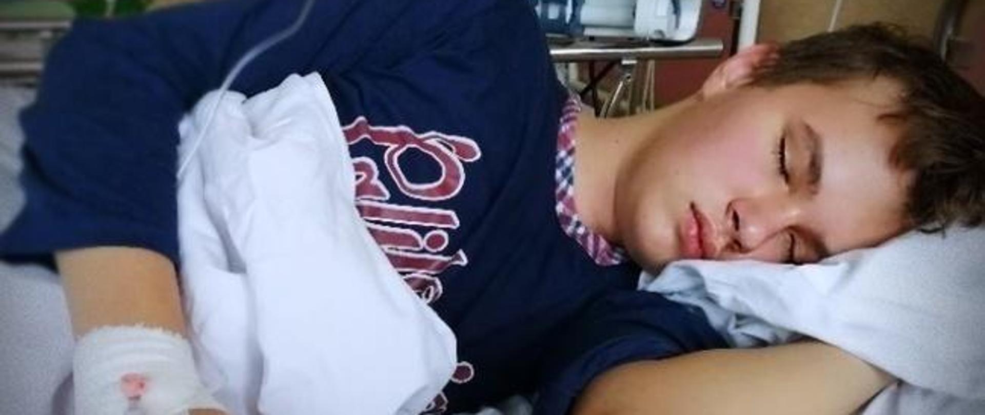 Mateusz leży lewym bokiem na łóżku szpitalnym, chłopiec ma zamknięte oczy, w tle aparatura szpitalna.