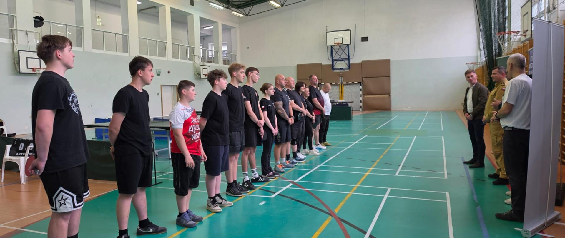 To zdjęcie przedstawia oficjalne rozpoczęcie lub zakończenie wydarzenia sportowego, prawdopodobnie turnieju tenisa stołowego, odbywającego się w hali sportowej.
Główne elementy zdjęcia:
Uczestnicy: Po lewej stronie, wzdłuż linii boiska, stoi grupa kilkunastu osób ustawionych w szeregu. Większość z nich ubrana jest w sportowe, ciemne stroje (głównie czarne koszulki i spodenki). Widać tu zróżnicowanie wiekowe – od młodzieży po osoby dorosłe.
Organizatorzy/Goście: Naprzeciwko szeregu, po prawej stronie zdjęcia, stoją trzej mężczyźni. Jeden z nich ma na sobie beżowy mundur służbowy (taki sam jak na poprzednim zdjęciu), co sugeruje obecność przedstawiciela Straży Pożarnej. Pozostali dwaj są w strojach cywilnych (jeden w garniturze/marynarce, drugi w białej koszulce).
Otoczenie: Akcja toczy się na dużej hali sportowej z charakterystyczną zieloną nawierzchnią pokrytą kolorowymi liniami wyznaczającymi boiska. W tle widać stoły do tenisa stołowego, kosze do koszykówki, drabinki gimnastyczne oraz antresolę z barierkami.
Konstrukcja hali: Sufit ma otwartą konstrukcję z metalowych kratownic, na których zamontowane są liczne lampy jarzeniowe oświetlające całe pomieszczenie.
Atmosfera:
Scena ma charakter formalny – uczestnicy stoją w postawie wyprostowanej, słuchając przemówienia osób stojących naprzeciwko. Obecność munduru oraz oficjeli sugeruje, że jest to wydarzenie o randze lokalnej, być może organizowane przez Powiat Warszawski Zachodni lub lokalną jednostkę OSP.