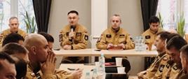 Strażak trzyma mikrofon siedząc za stołem obok niego siedzą inni funkcjonariusze Państwowej Straży Pożarnej przed nim naprzeciwko siebie siedzą strażacy.