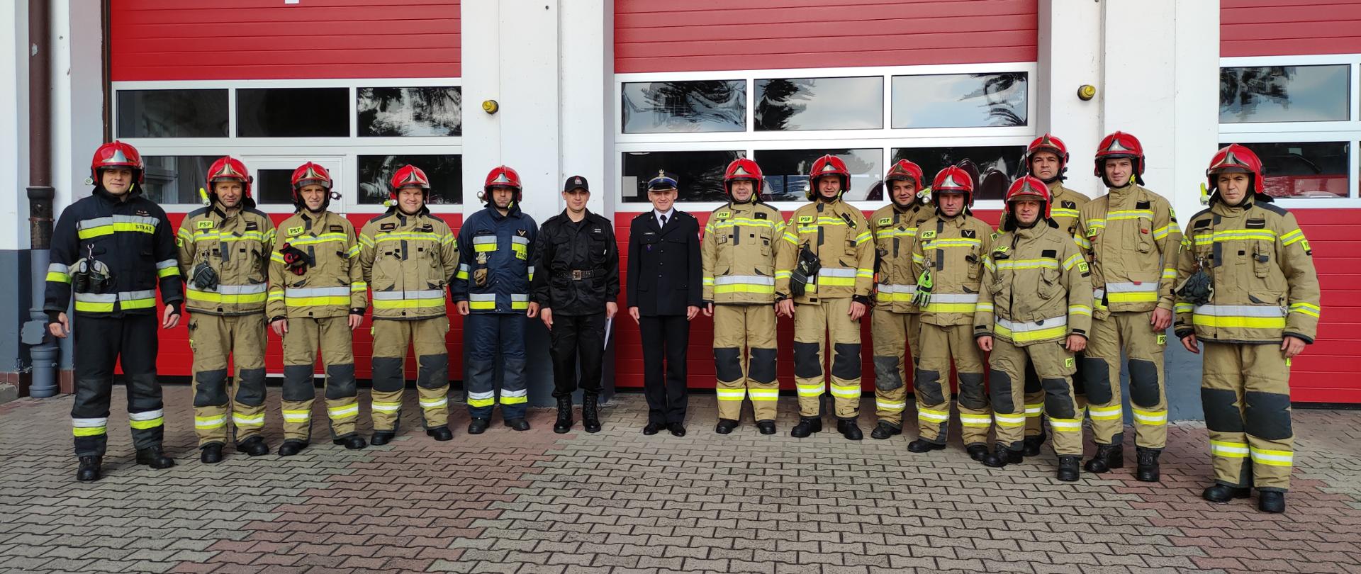 Komendant Powiatowy mianuje na dowódcę JRG podczas zmiany służby. Zdjęcie grupowe strażaków w ubraniach specjalnych i hełmach z dowódcą JRG i komendantem na tle budynku strażnicy.