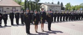 Na zdjęciu widzimy czterech strażaków stojących w szeregu. Strażacy ubrani są w galowe mundury koloru czarnego. Dwie osoby stojące w środku szeregu to kobiety w prawych rękach trzymają czerwone róże. W tle widzimy strażaków stojących w szeregu oraz budynki mieszkalne, zielone drzewa.