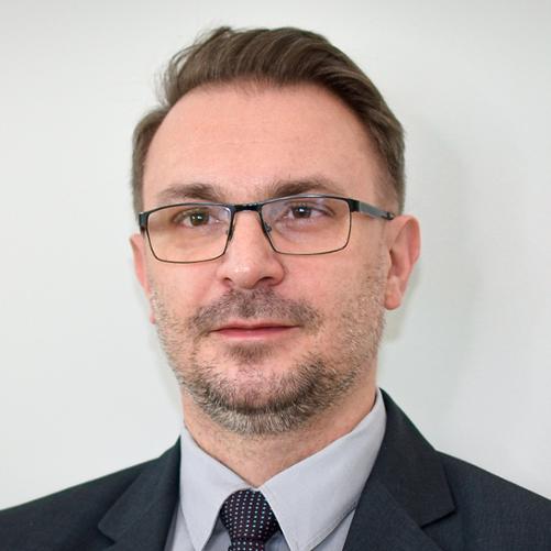 Mężczyzna w ciemnej marynarce, szarej koszuli i krawacie w biały wzorek, w okularach, z lekkim zarostem i krótkimi włosami.