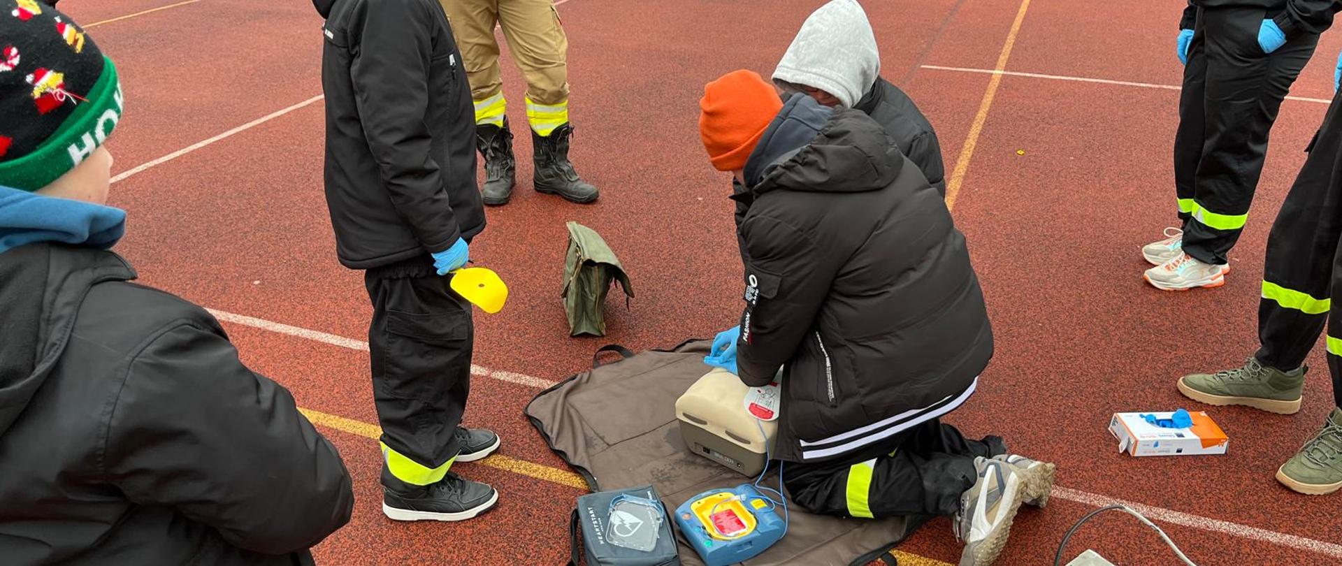 Młodzież podczas ćwiczeń z pierwszej pomocy na boisku używająca automatycznego defibrylatora zewnętrznego.