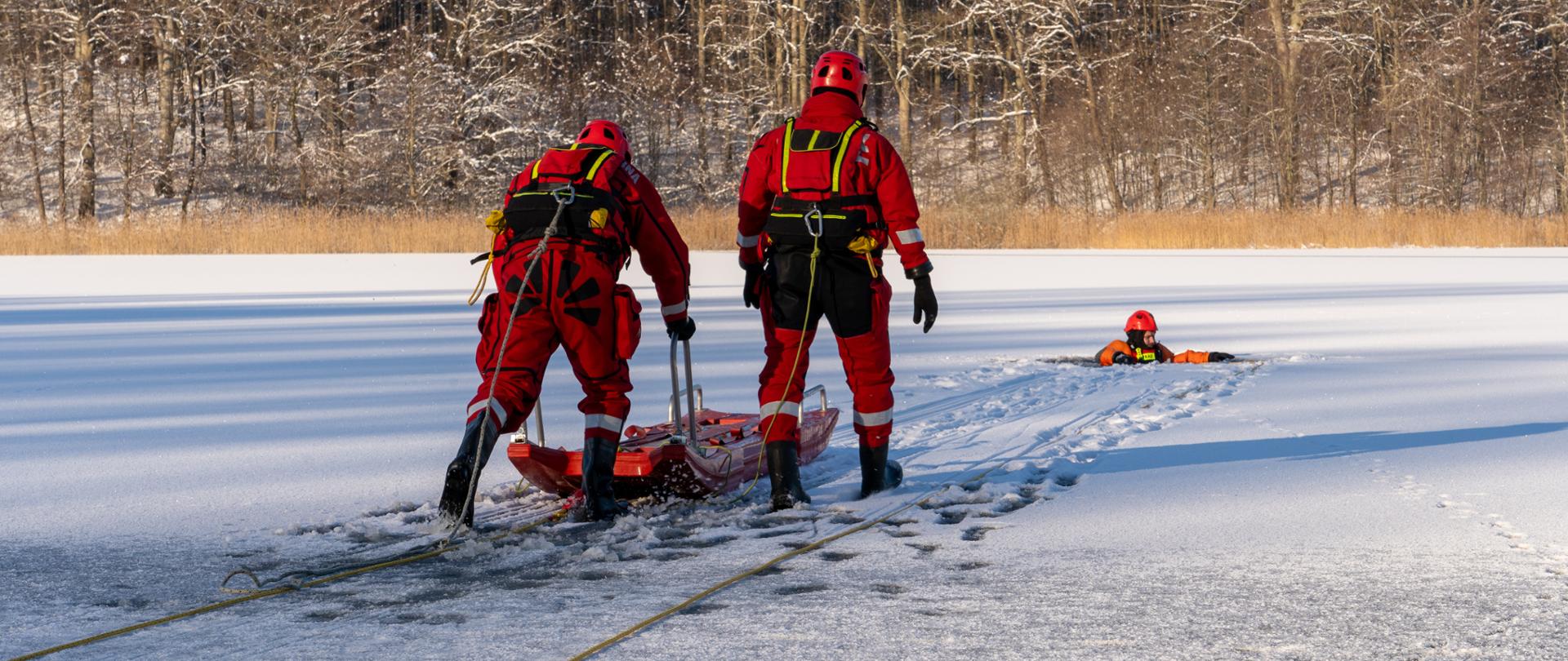 Strażacy w kombinezonach do pracy w wodzie z saniami wodno-lodowymi podchodzą do pozoranta znajdującego się w przerębli na zamarzniętym jeziorze 
