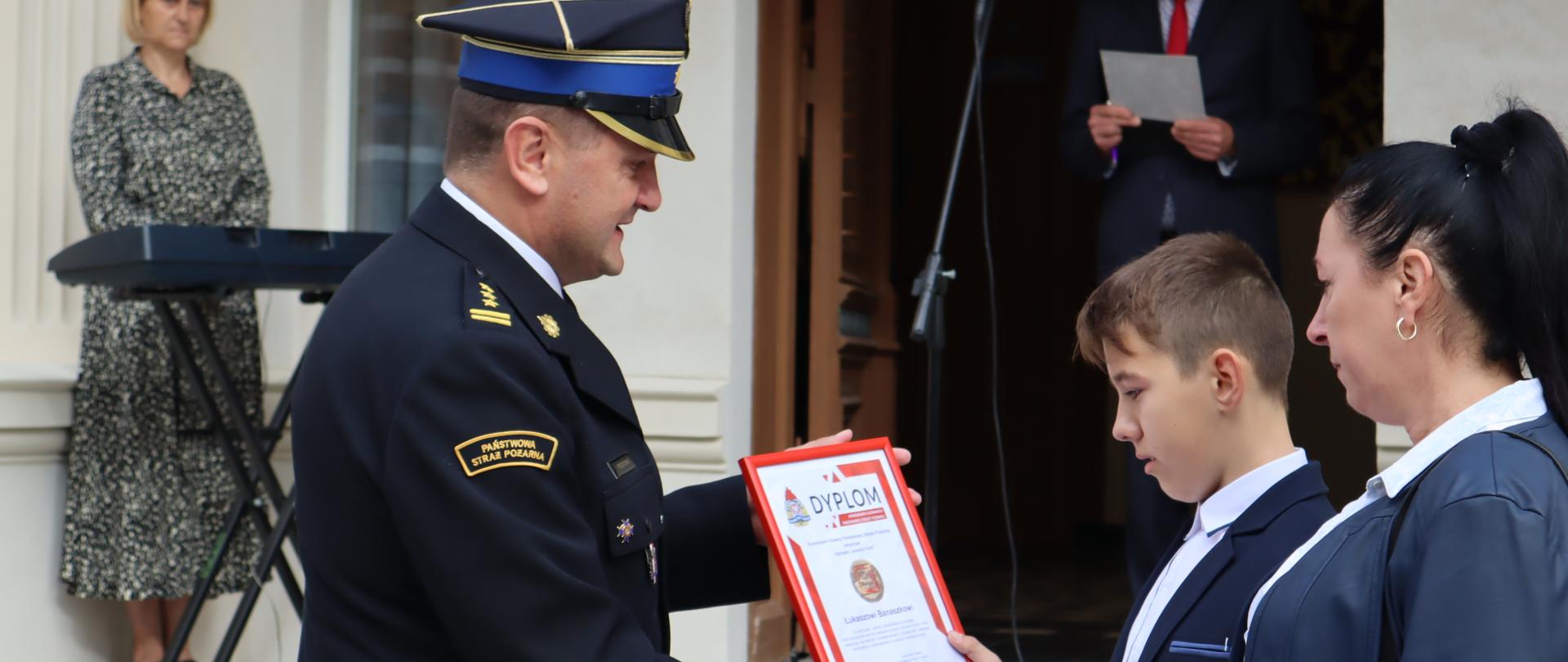 Komendant miejski PSP wręcza chłopcu odznakę i dyplom komendanta główngo PSP. Obok stoi mama chłopca, w tle budynek szkoły i dwie osoby z obsługi.